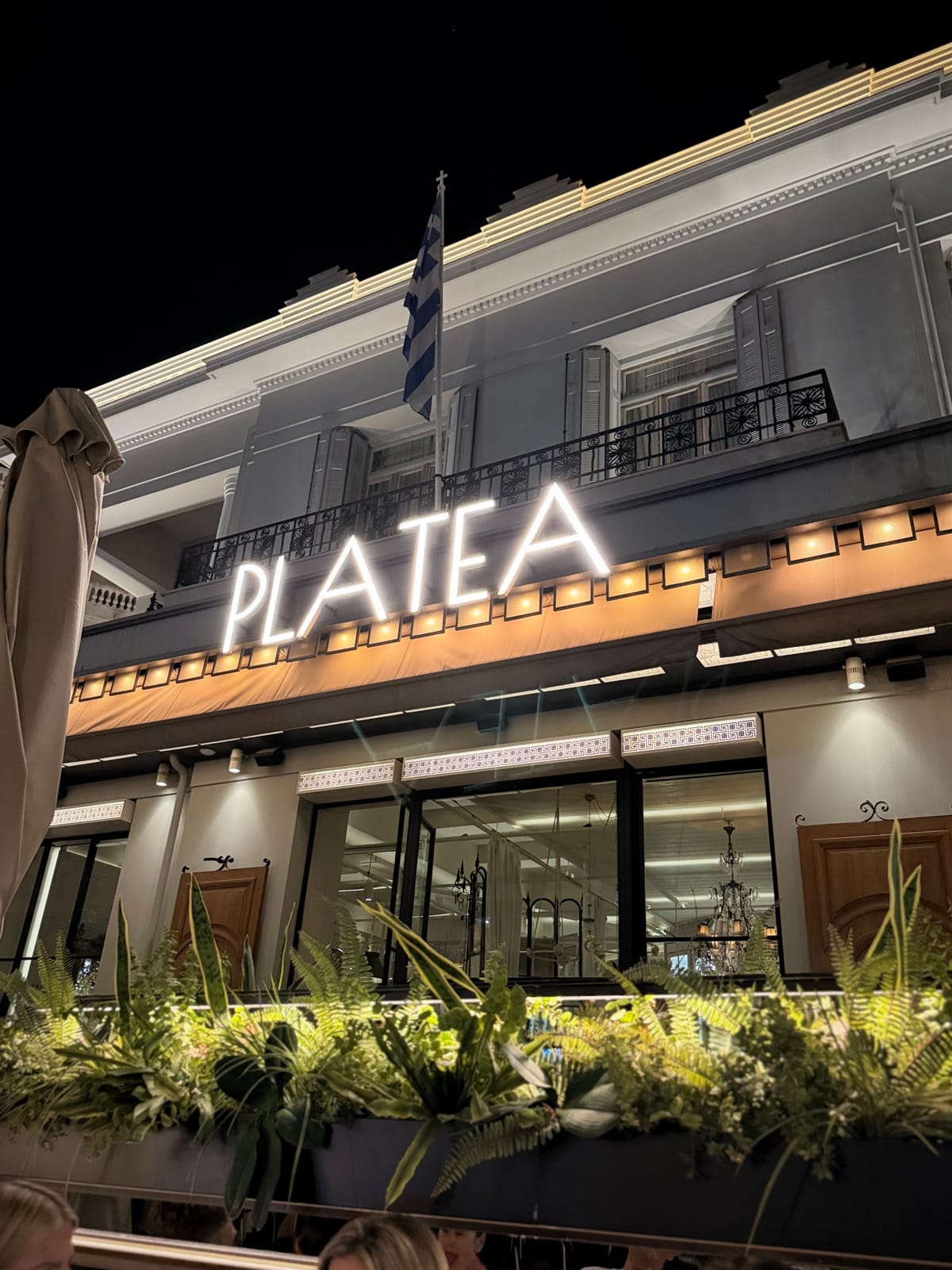 The illuminated neoclassical facade of a Kalamata restaurant at night, a Greek flag over the balcony and planted ferns along the terrace rail