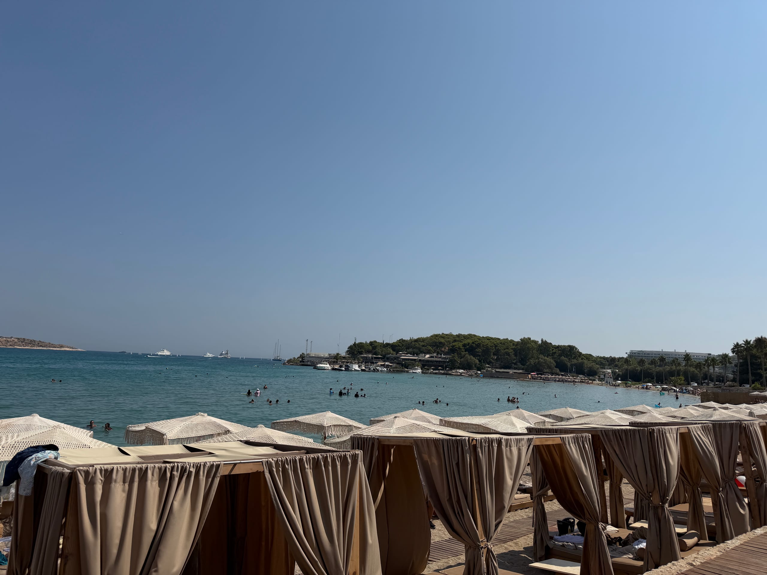 Cabanas and fringed umbrellas at a Glyfada beach club, swimmers in the shallows and the wooded point beyond