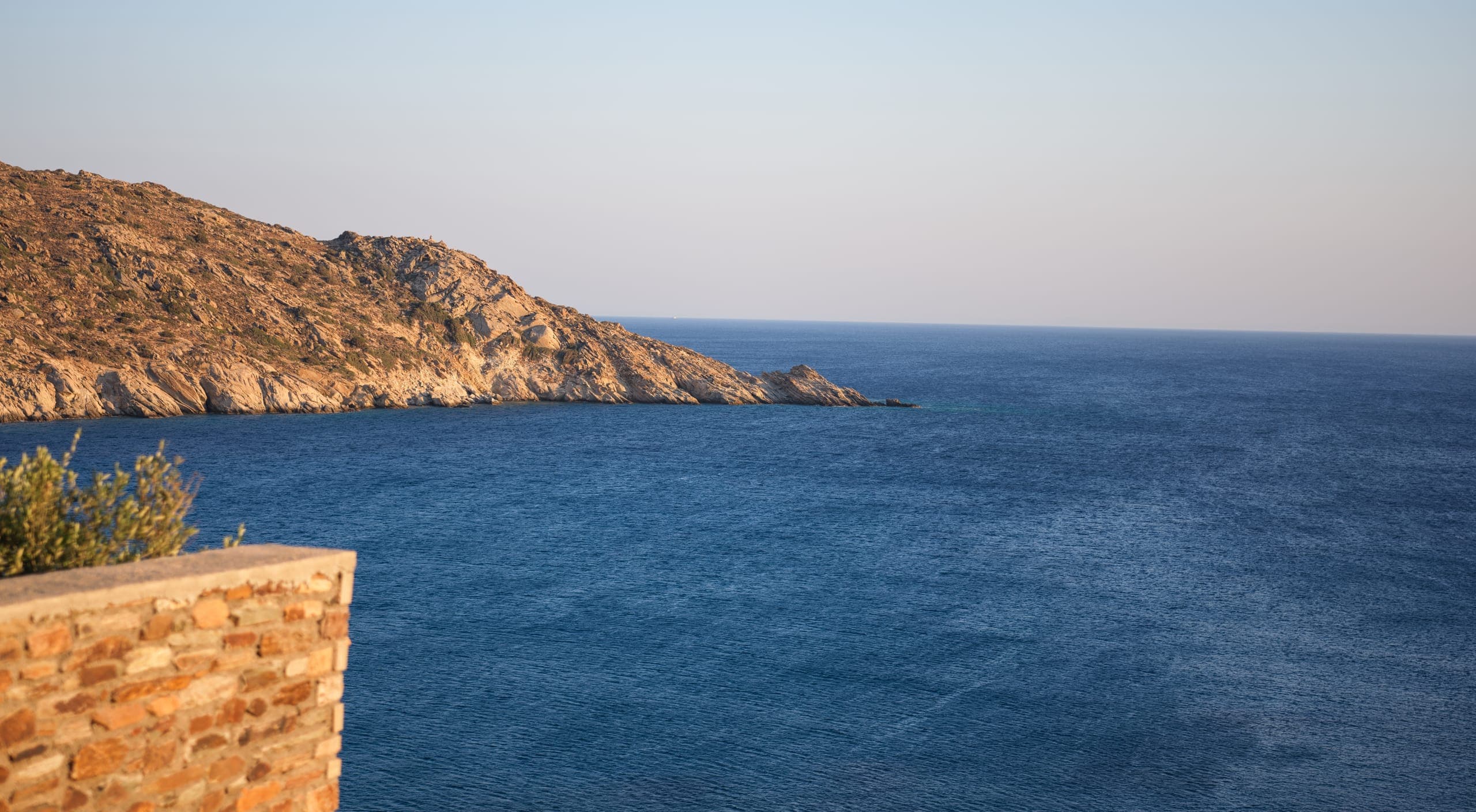 Golden-hour light on a rocky headland and calm deep-blue sea off Ios, seen past the corner of a dry-stone terrace wall