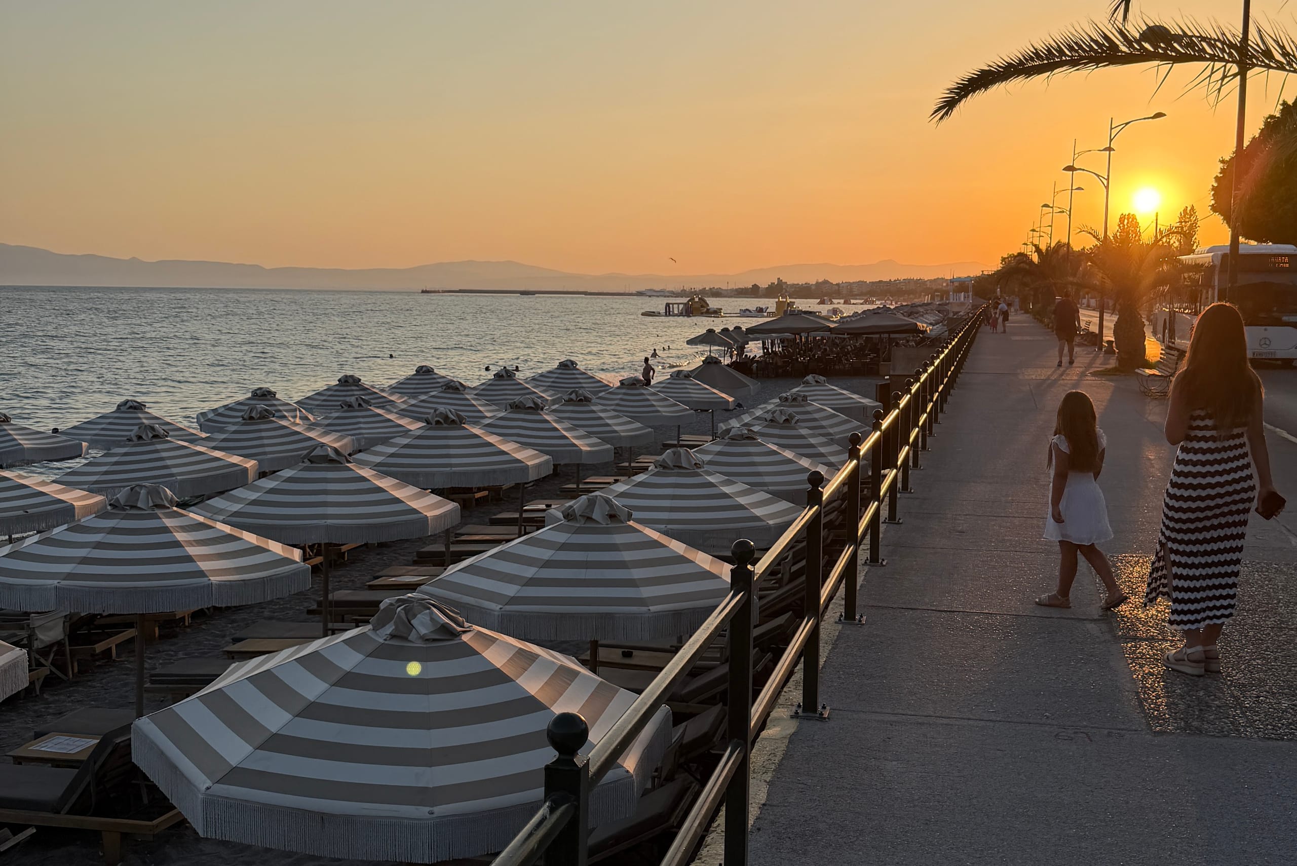 The Kalamata seafront at sunset — rows of fringed grey-and-white beach umbrellas along the sand, the Messenian Gulf beyond, and the promenade running toward the low sun