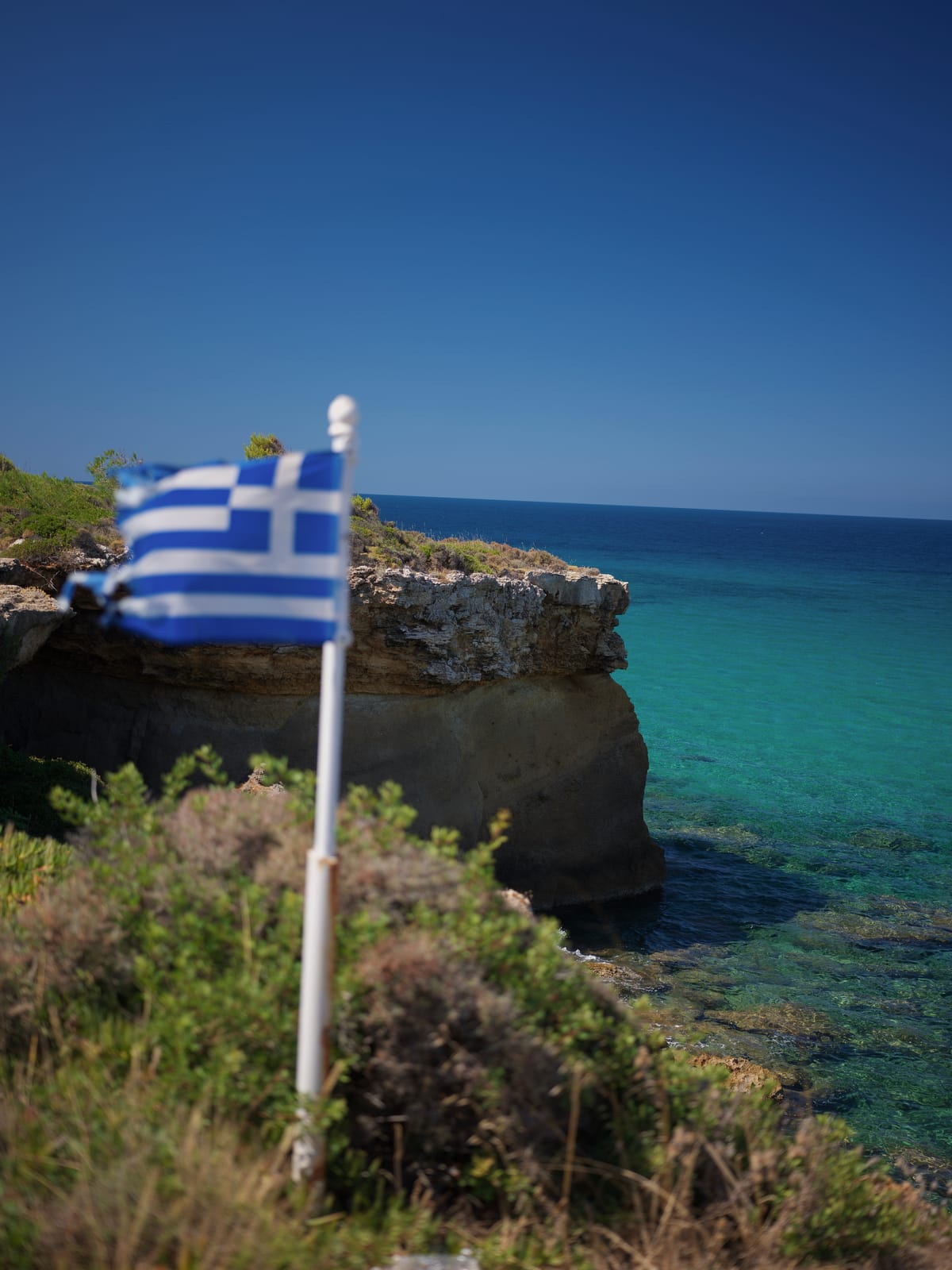A weathered Greek flag on a white pole above clear turquoise water on the Kefalonia coast