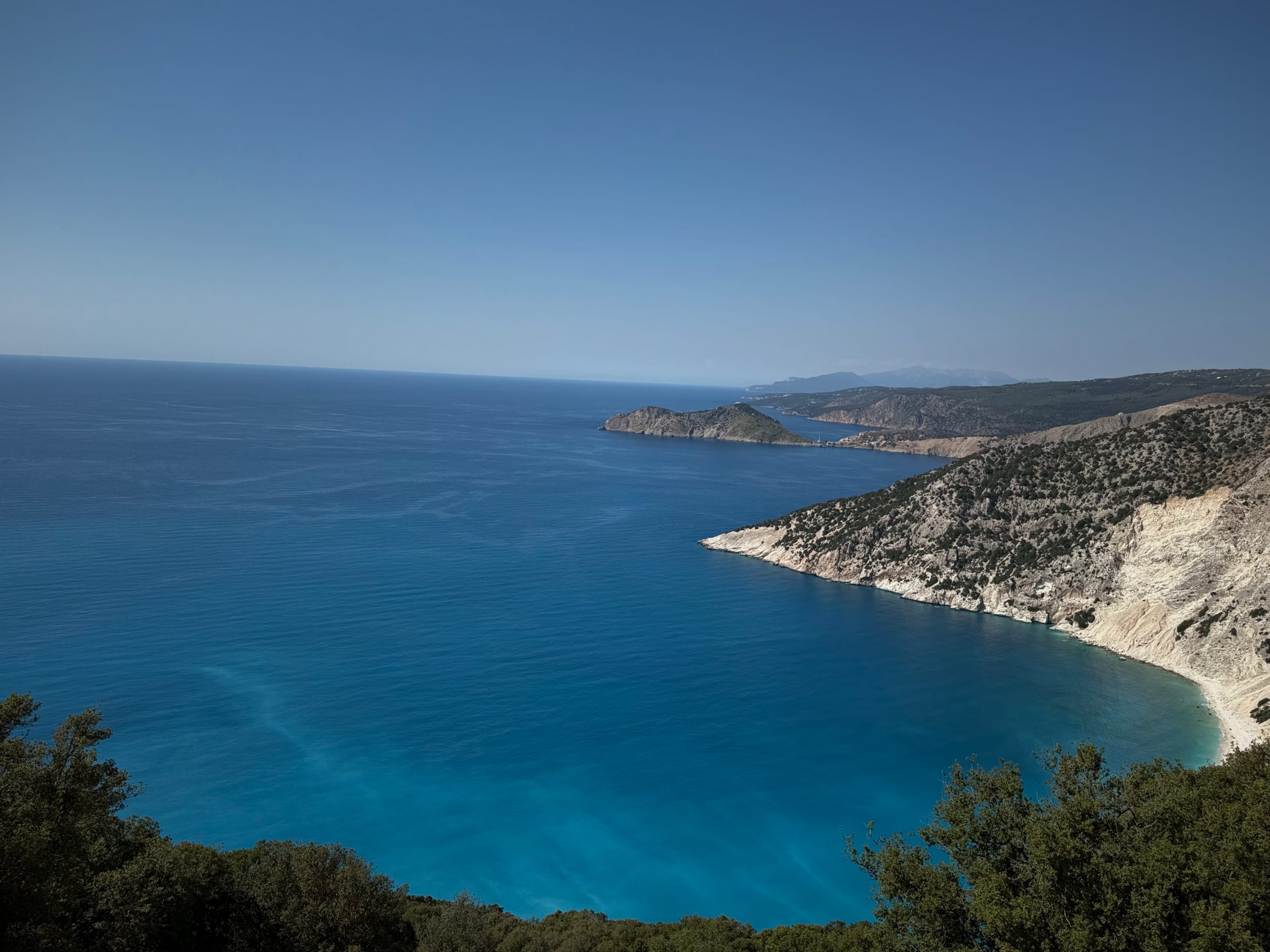 Myrtos bay from the overlook above — white limestone cliffs dropping into deep turquoise water, the Assos headland beyond
