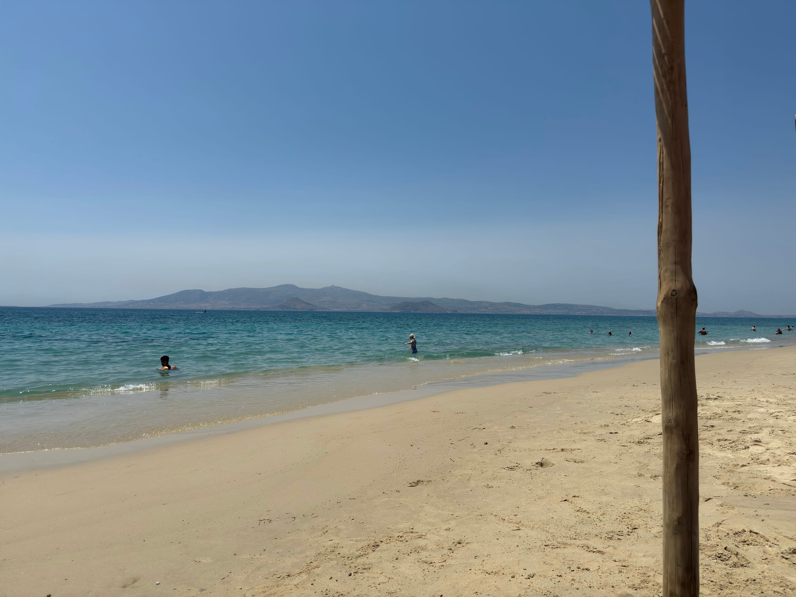 A broad empty stretch of pale sand on Naxos meeting shallow turquoise water, a long island ridge low on the horizon across the strait