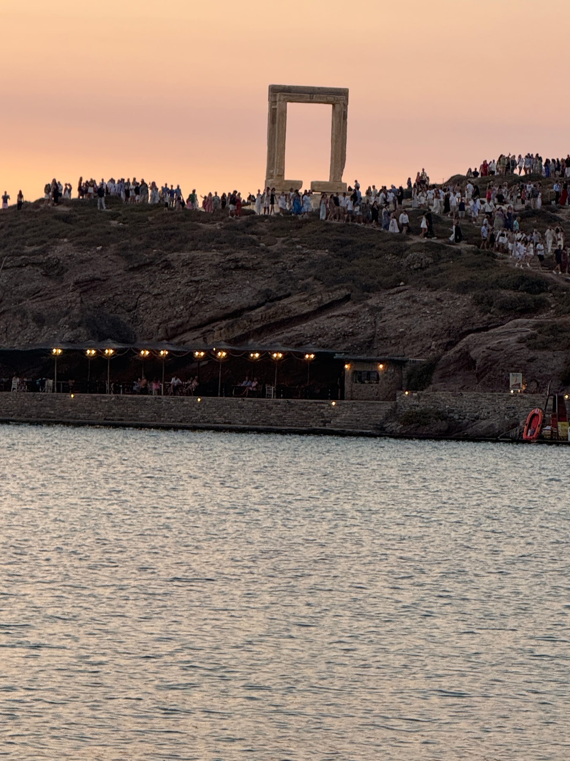 The Portara — a freestanding marble doorframe on the islet above Naxos harbour — silhouetted against a pink sunset, with a lit waterfront taverna on the quay below