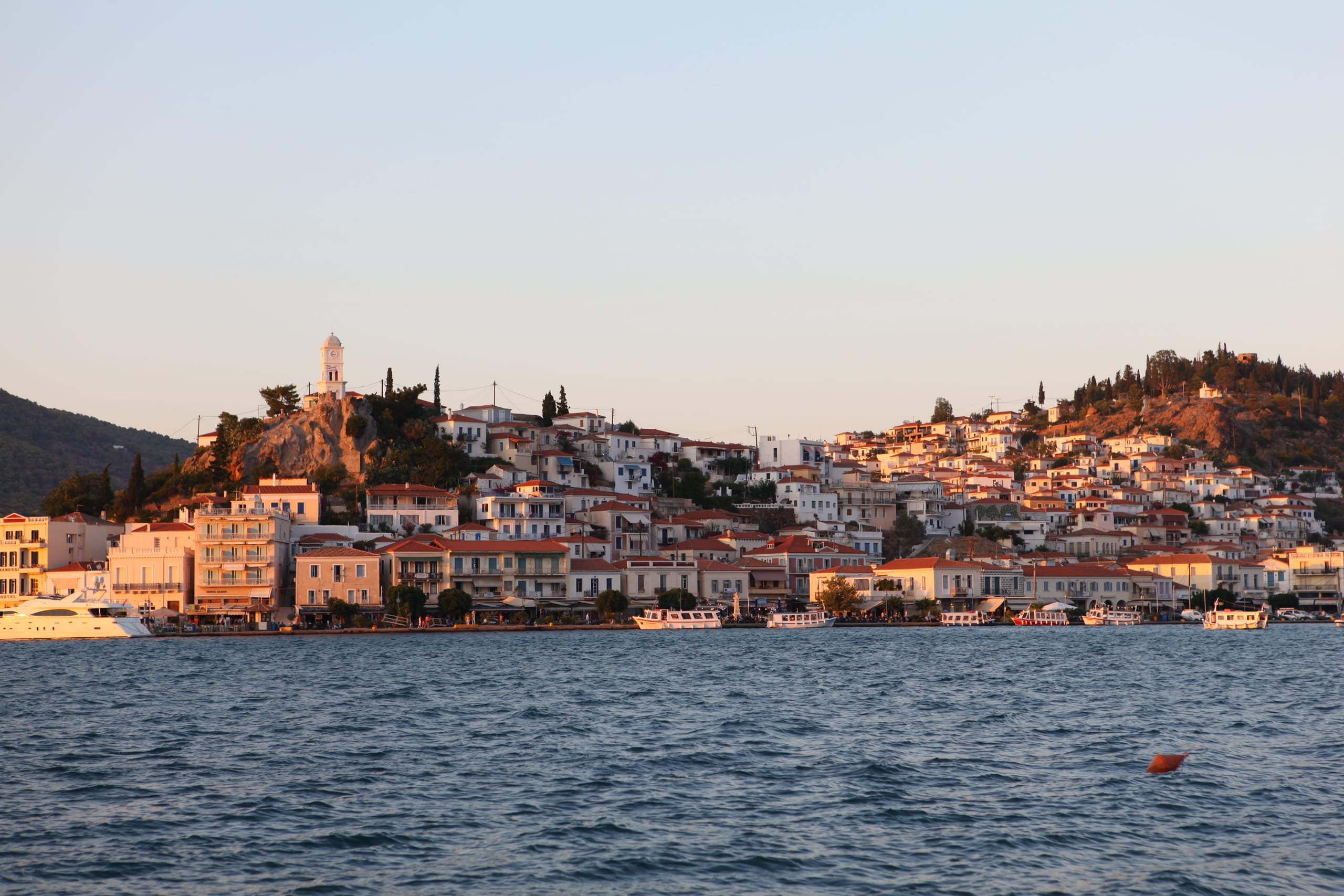 Poros town from the channel at low sun — tiered houses climbing to the clock tower, the waterfront and moored boats along the quay