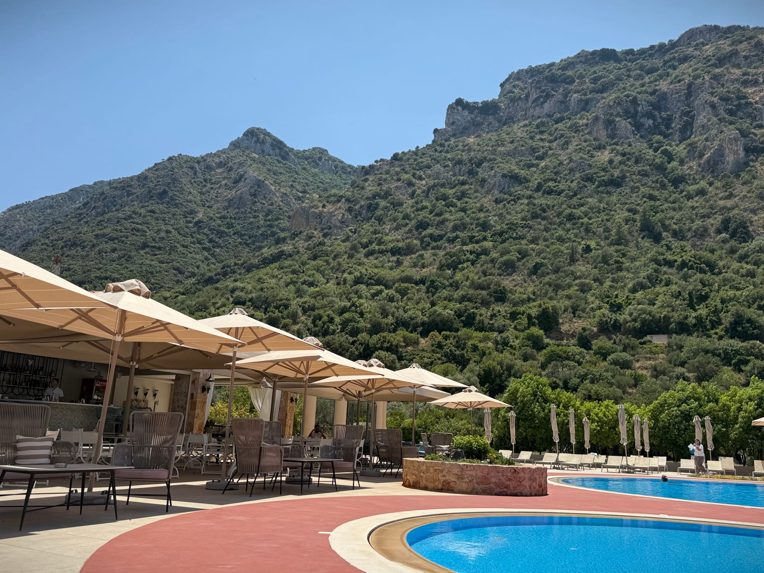 A hotel pool terrace in the Sparti area — cream square parasols and a shaded bar beside two blue pools, a scrub-covered limestone ridge rising behind