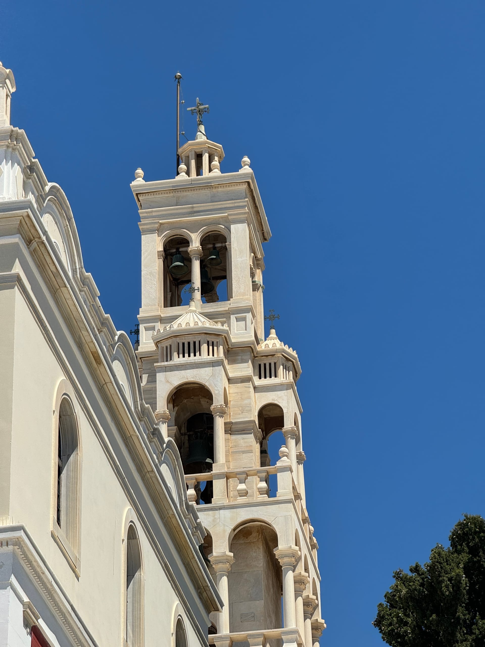 The white marble bell tower of Panagia Evangelistria against a cloudless sky, tiers of arched openings holding its bells
